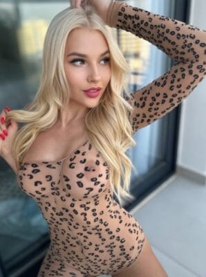 Blonde woman in a leopard-print bodysuit posing by a window, arm raised, looking to the side.