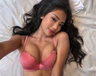 Woman with long dark hair lying on a white bed, wearing a pink bra and taking a selfie against the camera light.