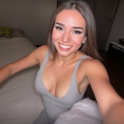 Smiling young woman taking a selfie in a bedroom, wearing a light gray tank top.