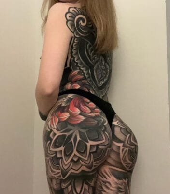 Side view of a person wearing a black thong, showcasing bold black-and-gray floral and mandala tattoos across the hip and buttock area.