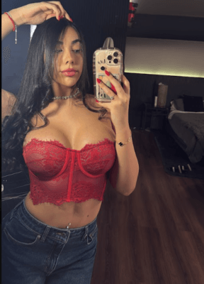 Selfie of a woman with long dark hair wearing a red lace bustier and denim jeans, taking a mirror photo in a bedroom.