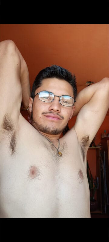 Man with short dark hair and glasses, shirtless, arms raised behind his head, looking at the camera in an indoor room with orange walls.