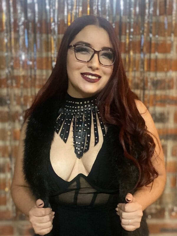 Smiling woman with dark hair and glasses, wearing a black outfit with a studded fringe collar and faux fur vest.