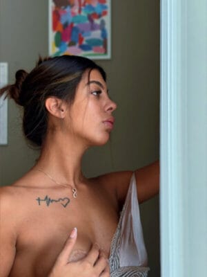 Side profile of a topless woman wearing a delicate necklace, with a heartbeat tattoo on her collarbone, standing by a doorway in a softly lit room.